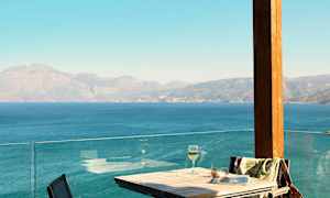 Bord og stoler på en terrasse med utsikt over havet og fjellene. (AI-generert tekst)