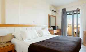 Et hotellrom med en dobbeltseng med hvite laken og et mørkt teppe. Rommet har også et skrivebord med speil og en balkong med utsikt. (AI-generert tekst)