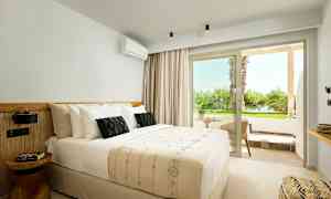 Interiør av et lyst hotellrom med en dobbeltseng og en liten sittegruppe utenfor med utsikt over grøntområder. (AI-generert tekst)