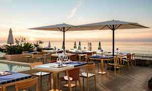 En utendørsrestaurant ved sjøen under solnedgangen med bord og stoler samt store parasoller. (AI-generert tekst)