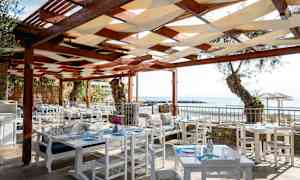 Uteservering med hvite bord og stoler under et tak av tre. Havutsikt i bakgrunnen. (AI-generert tekst)