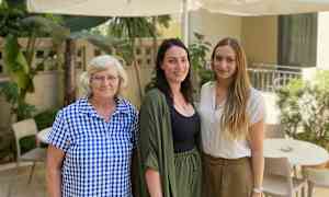 Tre personer står utendørs ved et bord og stoler under en parasoll. (AI-generert tekst)