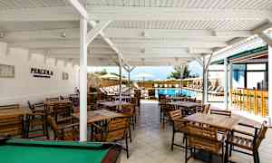 Uteservering med flere trebord og stoler under et hvitt tak nær et utendørs svømmebasseng. (AI-generert tekst)