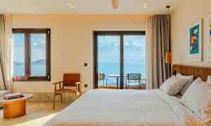 Et hotellrom med en oppredd seng, stoler og et bord ved et vindu med utsikt mot havet. (AI-generert tekst)