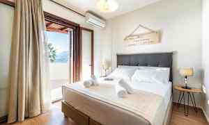 Et soverom med en dobbeltseng, dekorativ veggskilt, senge lamper og en åpen balkongdør som fører ut til en terrasse. (AI-generert tekst)