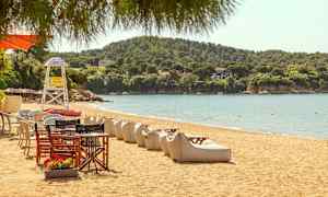 En sandstrand med solsenger og bord, med grønne åser i bakgrunnen. (AI-generert tekst)