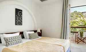 Rommet viser to enkeltsenger med gullfargede sengetepper og en dekorativ pute på hver seng. En dekorasjon henger over hodegavlen, og et vindu med utsikt over grøntområder og et hus ses til høyre. (AI-generert tekst)