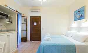 Et hotellrom med to senger kledd i blå sengetøy, en tredør, et lite kjøkken med vask og et maleri på veggen. (AI-generert tekst)