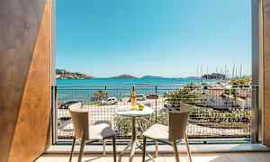 Utsikt fra en balkong med to stoler og et bord, mot havet og en småbåthavn. (AI-generert tekst)