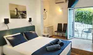 Et hotellrom med en oppredd seng og mørkeblå sengetøy, et åpent vindu mot balkongen, og møbler inkludert to stoler og en kommode. (AI-generert tekst)