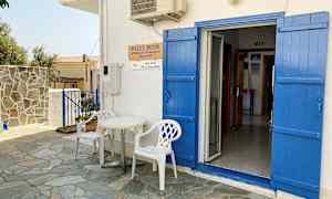 En hvit bygning med blå dører og skodder. Foran døren står to hvite plaststoler og et lite bord. (AI-generert tekst)
