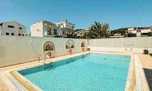 Et rektangulært svømmebasseng omkranset av en lys uteplass og en lav hvit mur, med bygninger og trær i bakgrunnen. (AI-generert tekst)