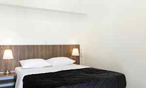 Et soverom med en dobbeltseng, hvitt sengetøy og et mørkt sengeteppe. To sengelamper står på nattbordene ved siden av sengen. (AI-generert tekst)