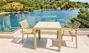 To beige stoler og et bord på en terrasse med glassrekkverk og utsikt over en bukt med båter og grøntområder. (AI-generert tekst)