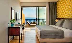 Hotellrom med dobbeltseng, skrivebord og havutsikt gjennom glasspanel. (AI-generert tekst)