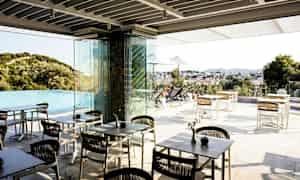 En utendørs terrasse med bord og stoler nær et infinitybasseng, med utsikt over et landskap og en by. (AI-generert tekst)