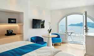 Hotellrom med lyse hvite vegger, møblert med en seng, bord og stoler, og en balkong med utsikt over havet og en øy. (AI-generert tekst)