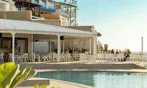 En utendørs restaurant med hvite møbler ved siden av et basseng, med noen personer som sitter ved bordene. Bakgrunnen består av bygninger og en lys himmel. (AI-generert tekst)