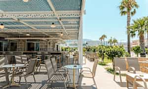 En utendørs terrasse med bord og stoler under et overdekket tak, omgitt av grøntområder og palmer. (AI-generert tekst)