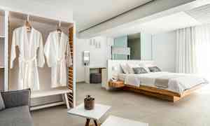 Bilde av et elegant hotellrom med en stor seng, hvite badekåper som henger i et skap og lyse moderne møbler. (AI-generert tekst)