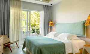 Hotellrom med dobbeltseng, blå sengklær og store vinduer som vender mot en frodig balkong. (AI-generert tekst)