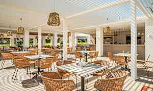 En utendørs restaurant med sitteplasser og bord, dekket med hvite pergolatak og dekorert med rottinglamper. Baren er synlig i bakgrunnen. (AI-generert tekst)