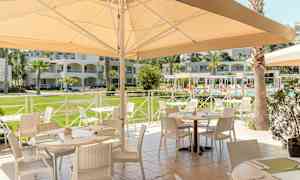 Utendørsrestaurant med møblerte bord nær et basseng og omkringliggende bygninger. (AI-generert tekst)