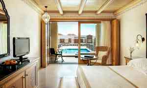 Hotellrom med trematerial inkludert en stor seng og en sittegruppe, samt en balkongdør som fører til et basseng. (AI-generert tekst)