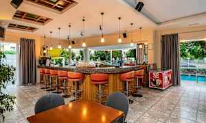 En bar med oransje barstoler og bardisk med belysning over. Utendørs vises trær og et basseng. (AI-generert tekst)