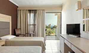 Hotellrom med to senger, TV, bord og stoler samt en balkong med utsikt mot grønne omgivelser og havet. (AI-generert tekst)