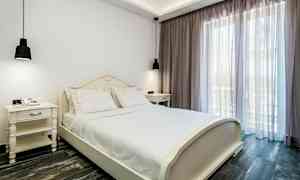 Et soverom med en hvit seng og hvite nattbord, ved siden av et stort vindu med lyse gardiner. (AI-generert tekst)