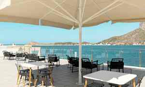 Utendørs serveringsområde med bord og stoler under en stor parasoll, med utsikt over havet og omkringliggende åser. (AI-generert tekst)