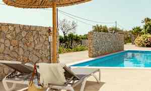 To solsenger med et håndkle og en stråveske ved siden av et basseng. En steinmur og palmer er i bakgrunnen. (AI-generert tekst)