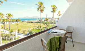 En balkong med bord, stoler og utsikt over en grønn hage med palmer og havet i bakgrunnen. (AI-generert tekst)