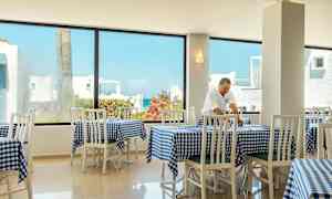 Lys restaurant med hvite stoler og rutete blå duker, samt store vinduer med utsikt over bygninger og grønne områder. (AI-generert tekst)