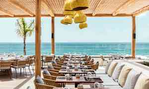 Restaurant med havutsikt, med bord og stoler i tre og rotting samt benker med puter under et beskyttende tak. (AI-generert tekst)