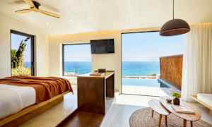 Et rom med utsikt over havet gjennom store vinduer, en seng er plassert til venstre og et bord med gjenstander er i forgrunnen. (AI-generert tekst)