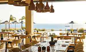 En restaurant med bord og stoler, med utsikt over et utendørs basseng, havet og parasoller. (AI-generert tekst)