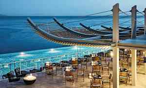 Restaurant med trestoler og bord ved et basseng, havet i bakgrunnen. (AI-generert tekst)