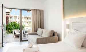 Et moderne hotellrom med en dobbeltseng, en grå sofa og en åpen glassdør som fører til en terrasse med stoler og grøntområder. (AI-generert tekst)