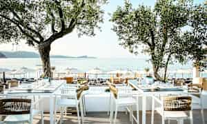 Utendørs servering med bord og stoler under trær, utsikt mot sjøen og parasoller på stranden. (AI-generert tekst)