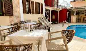 Bord og stoler ved et utendørs basseng, omgitt av bygninger med skodder. (AI-generert tekst)