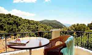 Utsikt fra en balkong med bord, drikke og en stol med et klede samt trær og fjell i bakgrunnen. (AI-generert tekst)