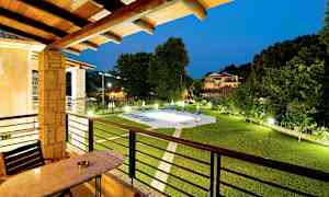 Utsikt fra en balkong med bord og stoler som viser en hage med et opplyst basseng og omkringliggende bygninger på kveldstid. (AI-generert tekst)