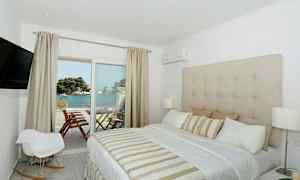 Et hotellrom med en stor seng, beige innredning og utsikt over havet fra en balkong. (AI-generert tekst)