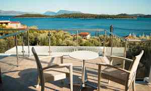 To stoler og et lite bord på en terrasse med glassrekkverk, utsikt mot havet og skogkledde øyer. (AI-generert tekst)