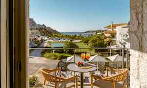 Utsikt fra en balkong med to stoler og et bord som har to glass med oransje drikke, mot en frodig terrasse og en havn. (AI-generert tekst)