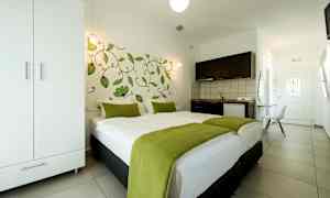 Et hotellrom med en dobbeltseng dekket med hvitt sengetøy og grønne tepper, et klesskap, en kjøkkenkrok og en dør i bakgrunnen. (AI-generert tekst)