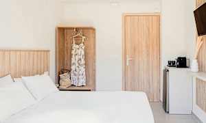 Et hotellrom med en hvit seng, et skap med en kjole og en veske, en dør og et lite kjøkkenområde med en kaffemaskin. (AI-generert tekst)