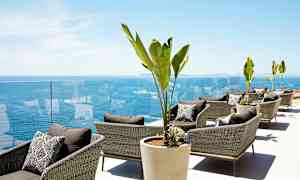 Loungeområde med rottingstoler, store potteplanter og utsikt over havet. (AI-generert tekst)